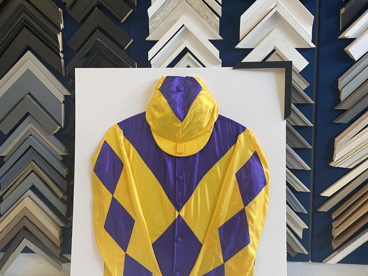 A set of racing silks float-mounted in a deep box frame, against the wall of moulding samples.